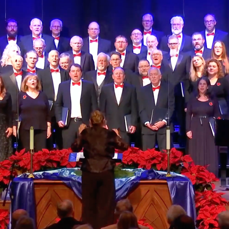 A choir of singers in black suits and red bowties performs behind a conductor and a row of red poinsettias against a dark background.