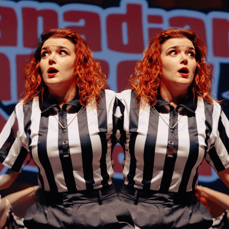 Two women with red hair wearing black and white striped referee shirts stand side-by-side in front of a blurred red and blue sign.