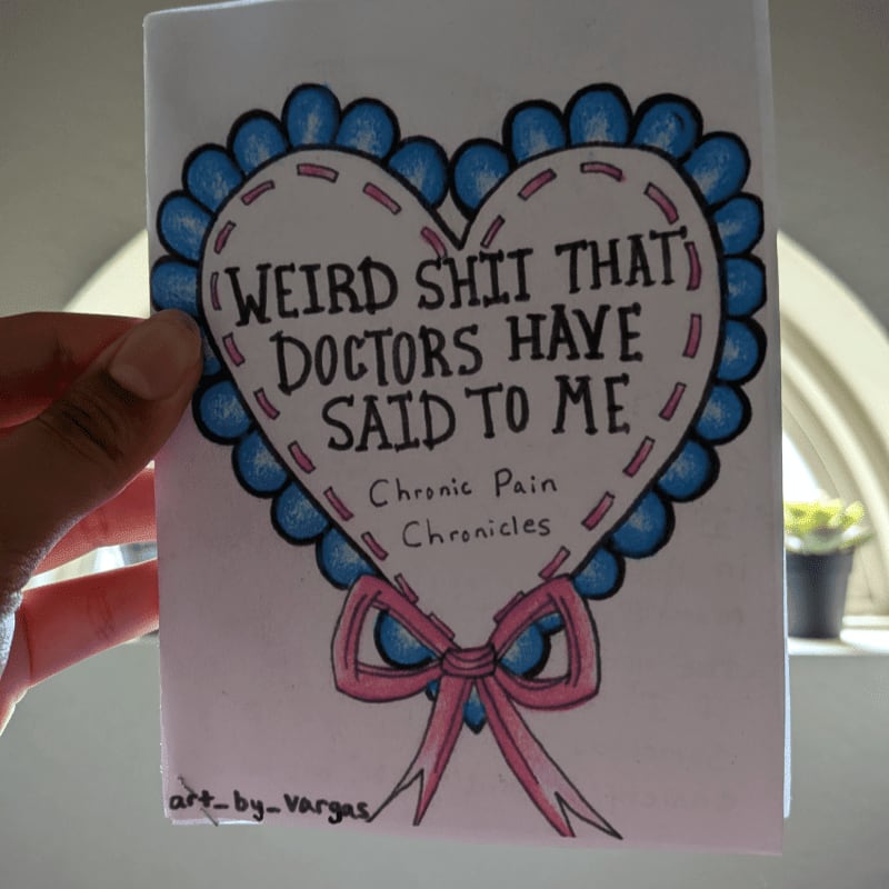 A hand holds a handmade card with a heart design that reads "Weird Shit That Doctors Have Said To Me: Chronic Pain Chronicles" against a blurred background of a window and a plant.