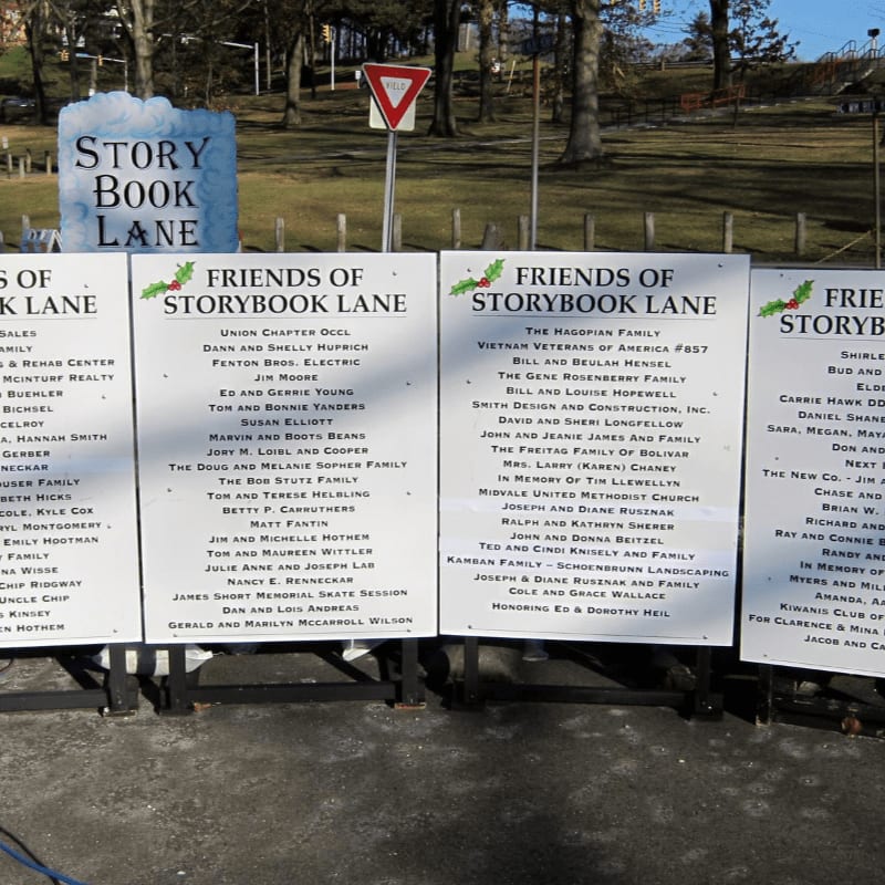 Four white plaques listing donors stand in the foreground against a background of a park with trees, a road, and a "Story Book Lane" sign.