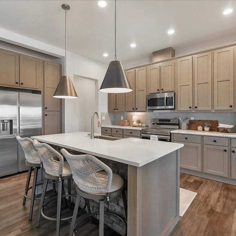 Three white woven barstools sit at a white kitchen island in the foreground, with light-colored cabinetry and stainless steel appliances in the background.