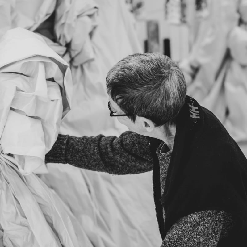 A woman in a dark sweater reaches toward a draped white sculpture in the foreground, with more fabric-covered forms blurred in the background.
