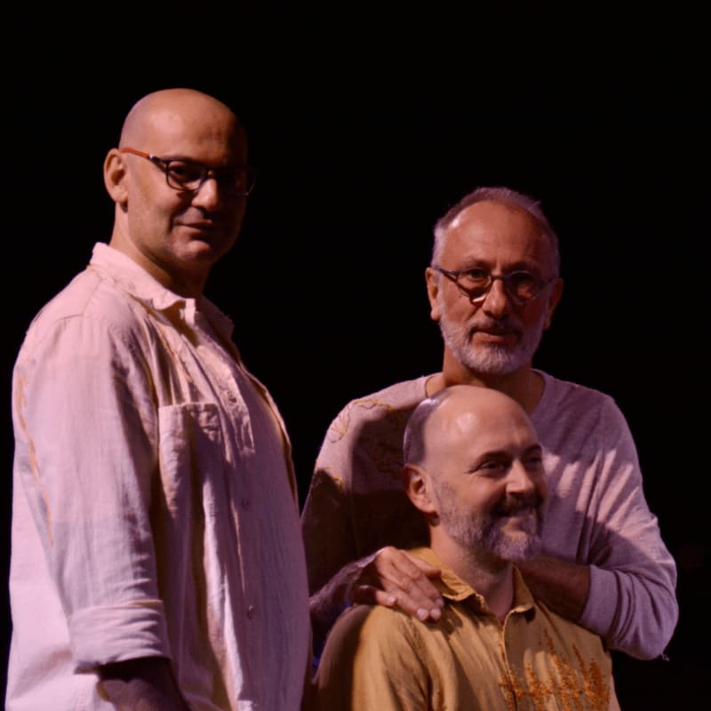 Three men wearing glasses and light-colored shirts stand together against a solid black background.