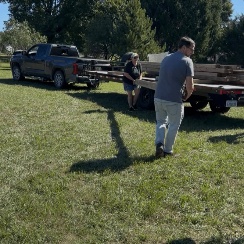 A man and woman stand in a grassy field next to a pickup truck towing a trailer loaded with lumber, with lush green trees in the background.