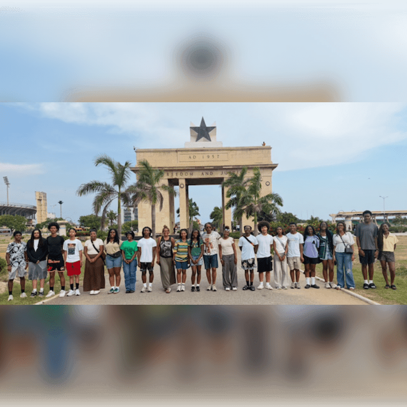 A group of young people stands in front of the Independence Arch in Accra, Ghana, with palm trees and a stadium visible in the background.