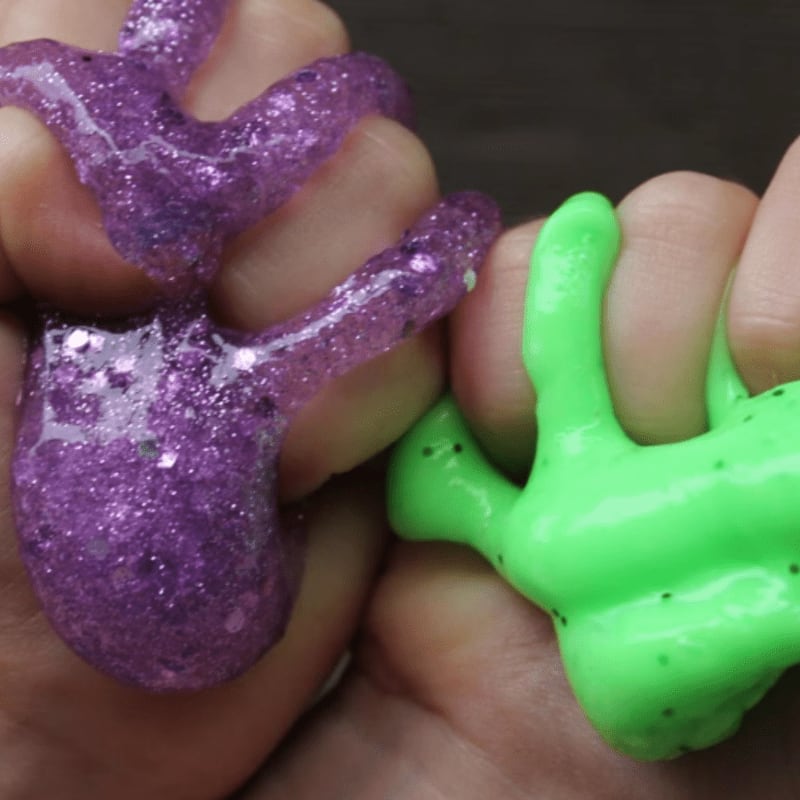 Hands hold glittery purple and neon green slime against a dark, blurred background.