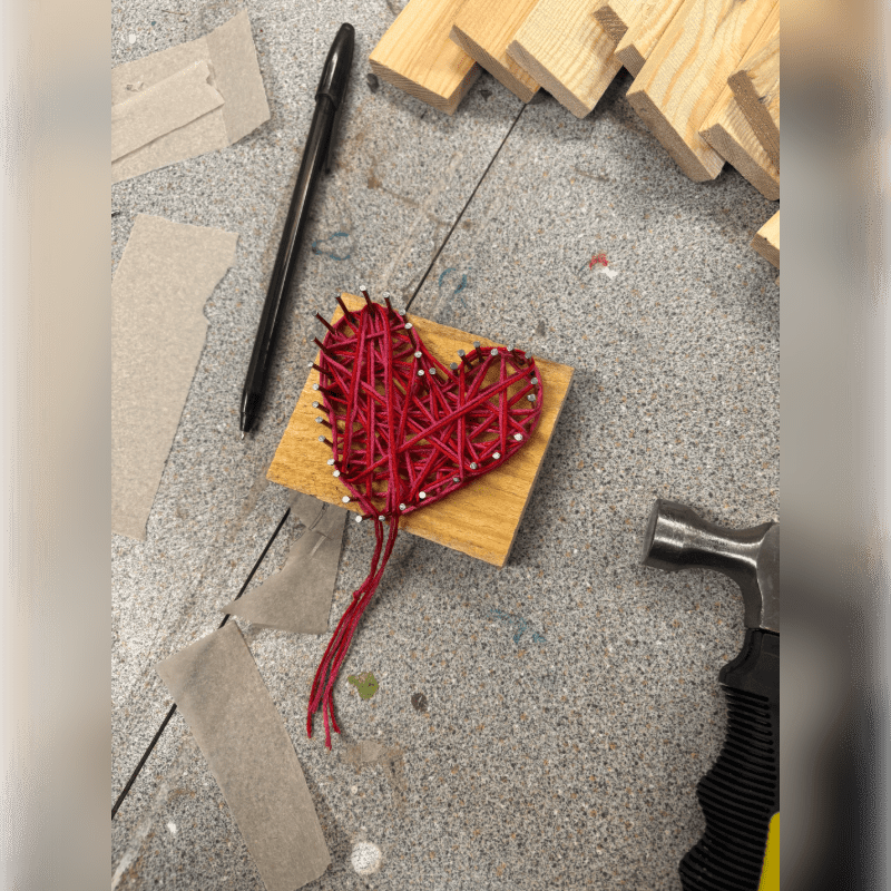 A red string heart pinned to a small wooden block sits in the foreground, surrounded by a pen, sandpaper, a hammer, and wooden slats on a grey textured surface.