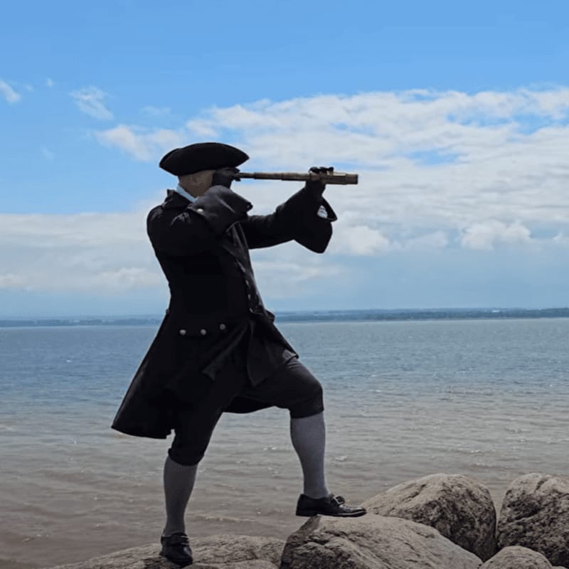 A man in colonial attire stands on rocks in the foreground, looking through a spyglass towards a vast body of water under a cloudy blue sky in the background.