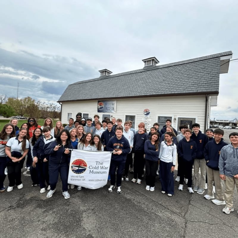 A large group of students poses for a photo in a parking lot in front of The Cold War Museum building.