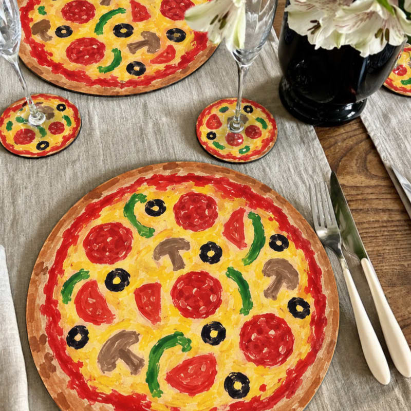 Two pizza-themed placemats and coasters sit on a grey linen tablecloth next to cutlery and a vase of white flowers.
