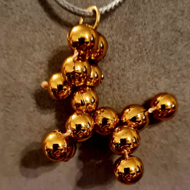 A gold-colored beaded pendant in a stylized shape hangs from a grey cord against a textured grey background.