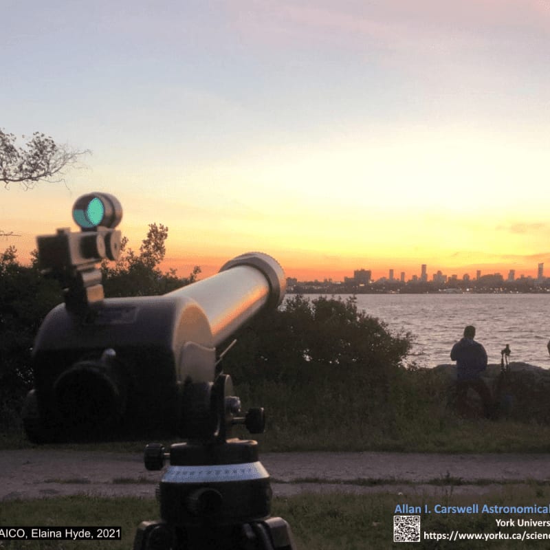 A telescope in the foreground points towards a distant city skyline across a body of water under a soft, colorful sunset sky.