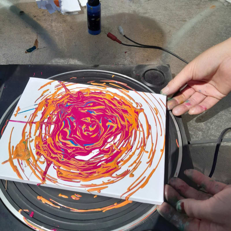 Hands hold a white canvas with a colorful swirl of paint on a rotating turntable, with art supplies scattered on the concrete background.