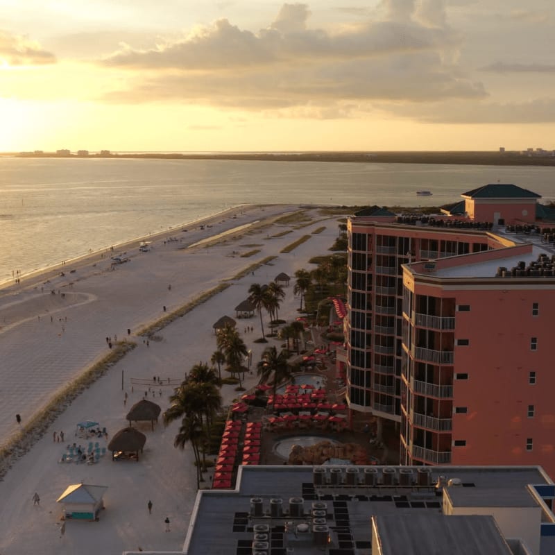 A tall pink resort overlooks a white sandy beach and calm ocean waters under a golden sunset sky.