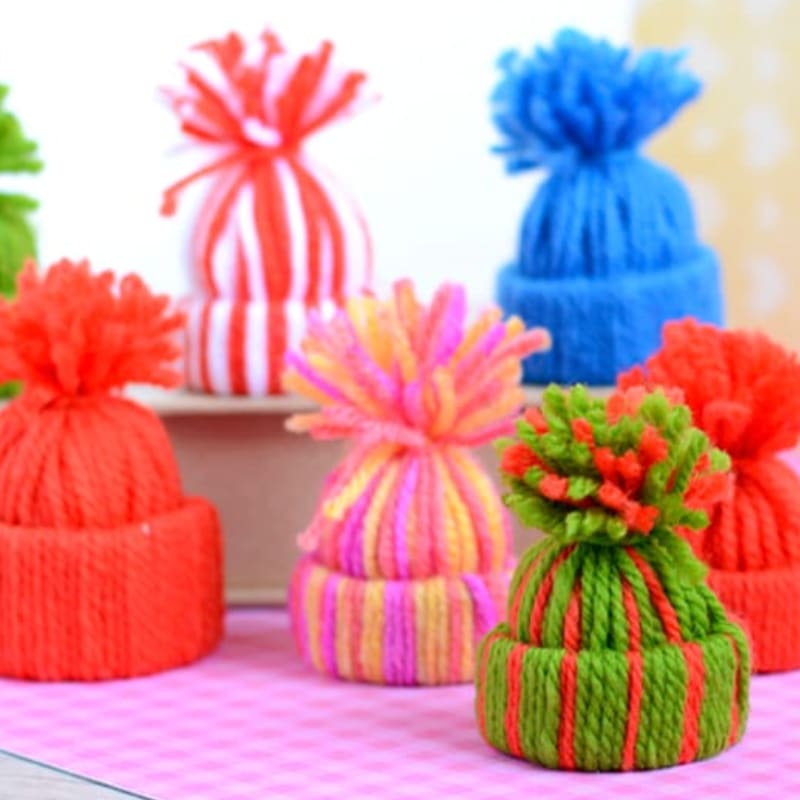 Several colorful knitted miniature hats with pom-poms sit on a pink checkered surface against a blurred white background.