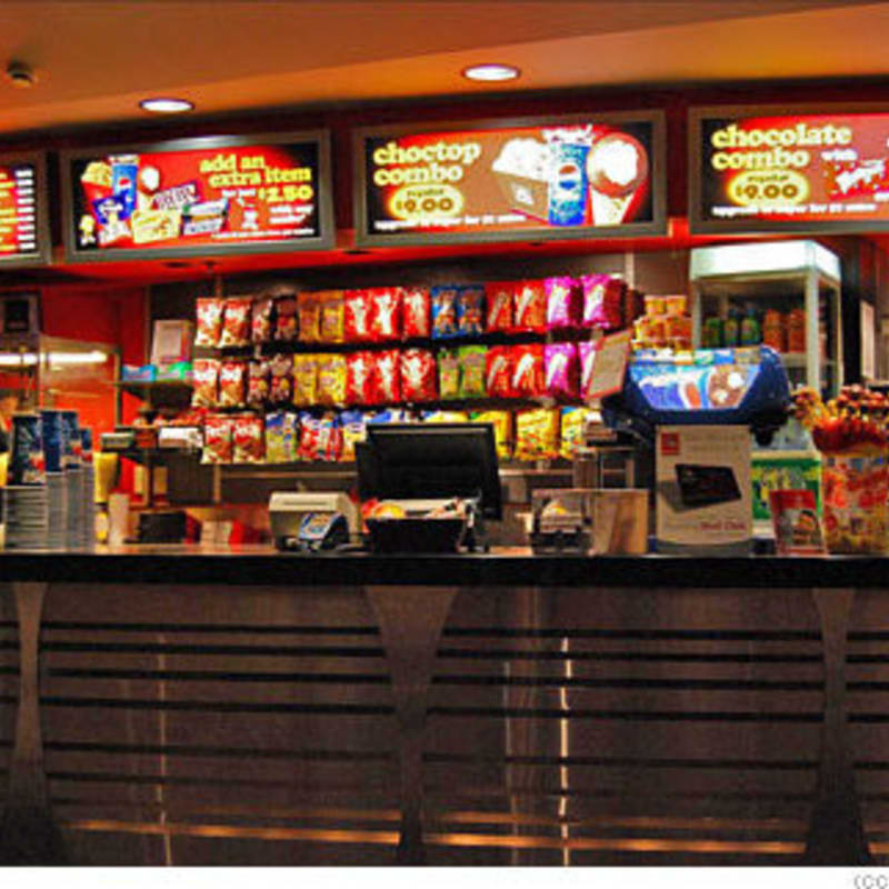 A sleek metal service counter sits in the foreground of a snack bar featuring shelves of chips and bright menu boards in the background.