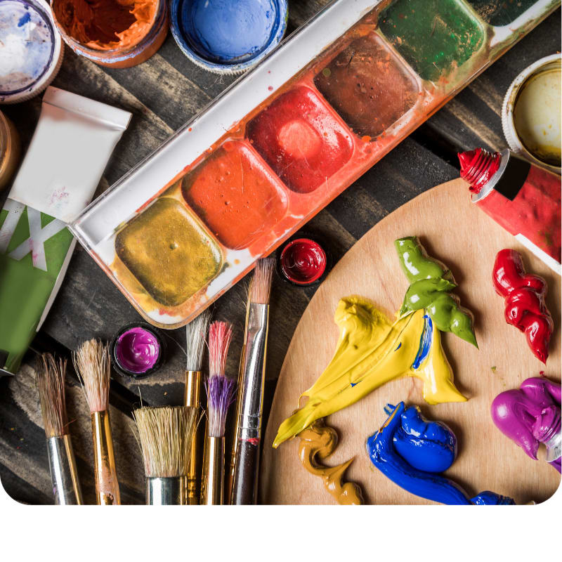Colorful paint tubes, brushes, and dollops of acrylic paint sit in the foreground on a wooden surface, with a multi-colored paint palette and small paint pots in the background.