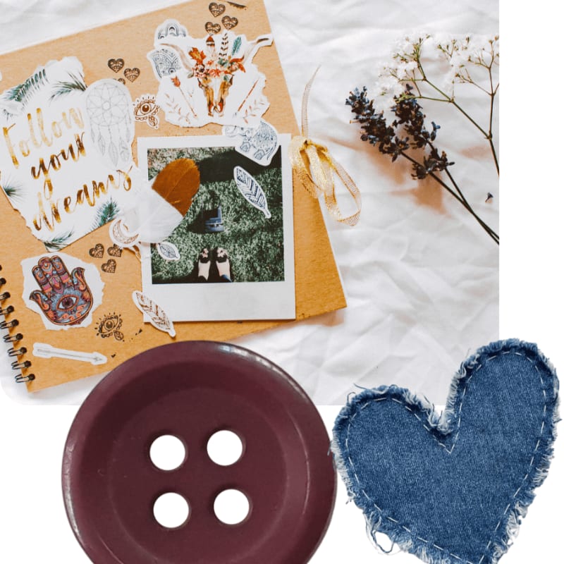 A large purple button and a denim heart sit in the foreground against a background of a scrapbook and dried flowers on a white cloth.