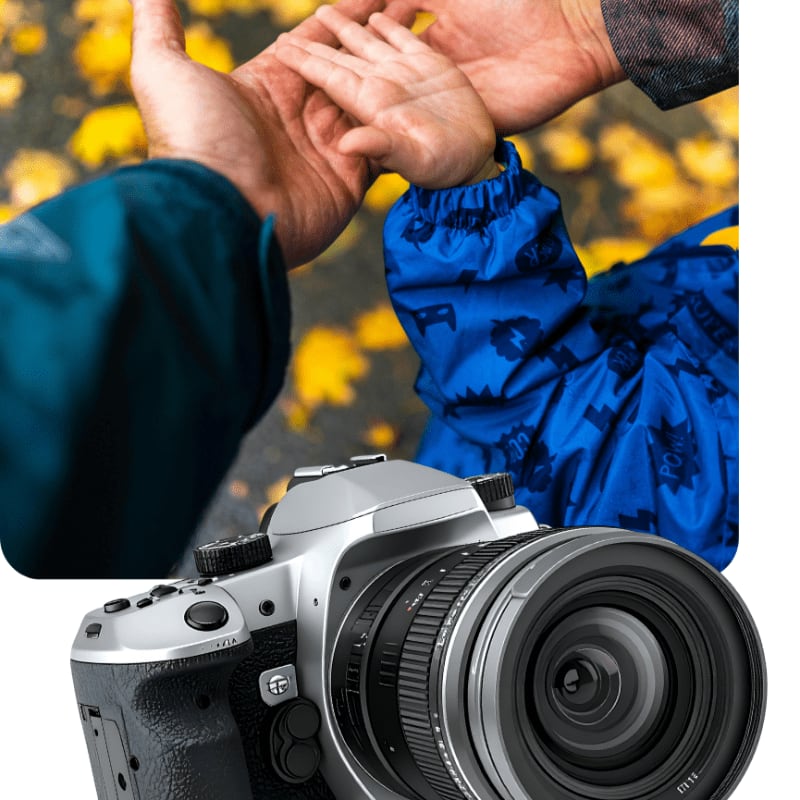 A silver digital camera sits in the foreground, while an adult and child hold hands against a background of fallen yellow leaves.