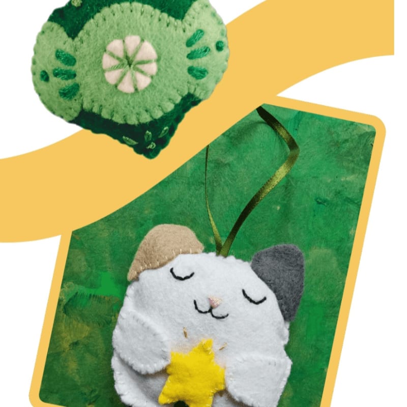 A white felt cat holding a yellow star sits in the foreground against a green background, with a green felt ornament positioned above on a yellow backdrop.