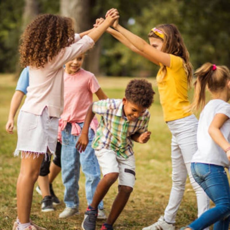 A group of children play together in a grassy field with blurred green trees in the background.