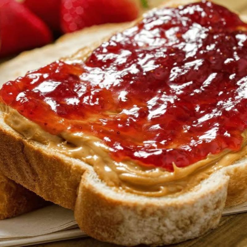 A slice of toast topped with peanut butter and red jam sits in the foreground with fresh strawberries in the background.
