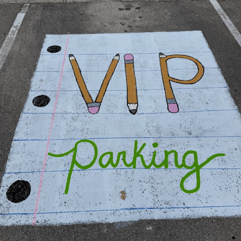 A parking space painted to look like a piece of notebook paper with "VIP Parking" written in pencils and green cursive is set against a grey asphalt background.