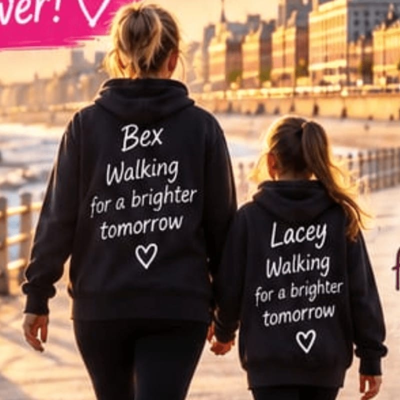 A woman and a young girl wearing matching black hoodies with personalized text walk along a seaside promenade with buildings in the background.