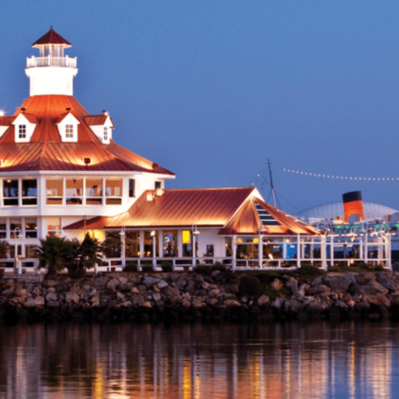 A brightly lit lighthouse-style building with orange roofs sits atop a rocky shoreline, reflecting in the calm water in the foreground under a deep blue twilight sky.