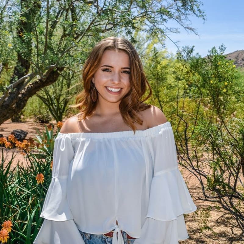 A smiling woman in a white off-the-shoulder top stands in the foreground against a background of green shrubs and trees under a clear blue sky.