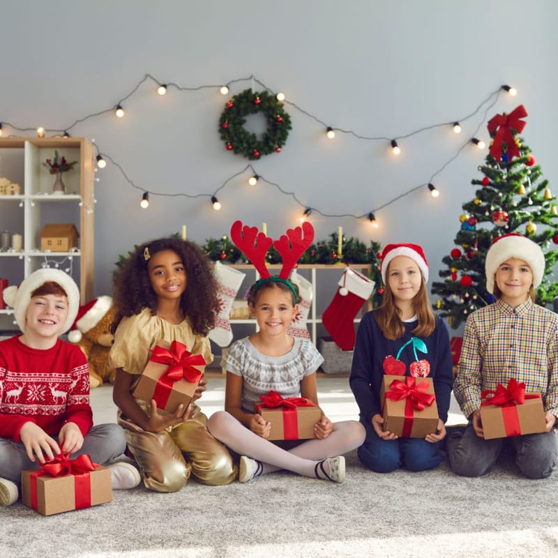 Five children wearing festive hats and holding wrapped gifts sit on a rug in front of a decorated Christmas tree and a wall with string lights and a wreath.