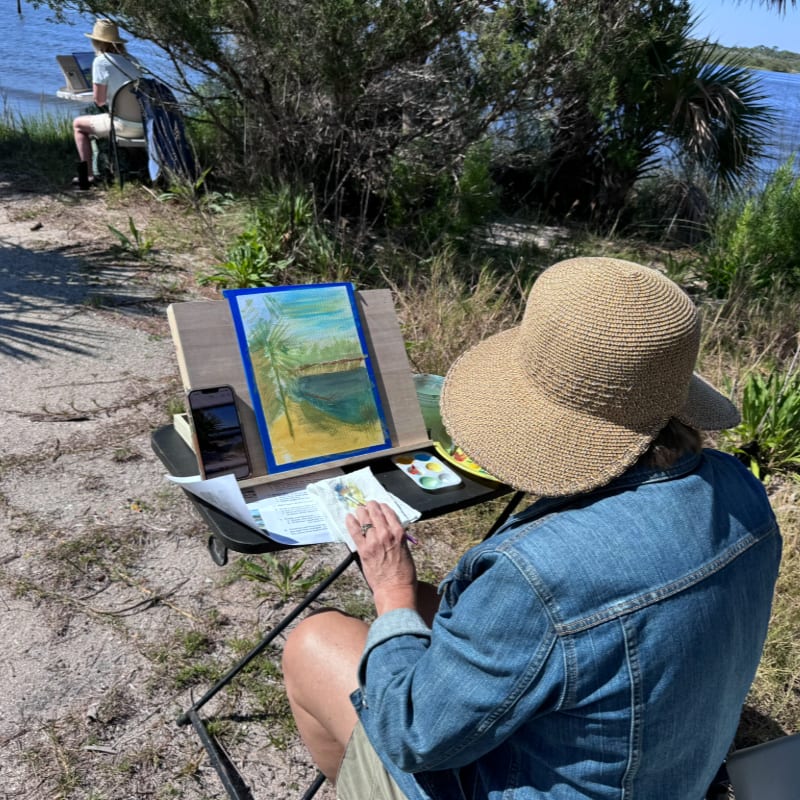 A person in a straw hat and denim jacket paints a landscape on an easel in the foreground, with another artist and a waterfront shoreline in the background.
