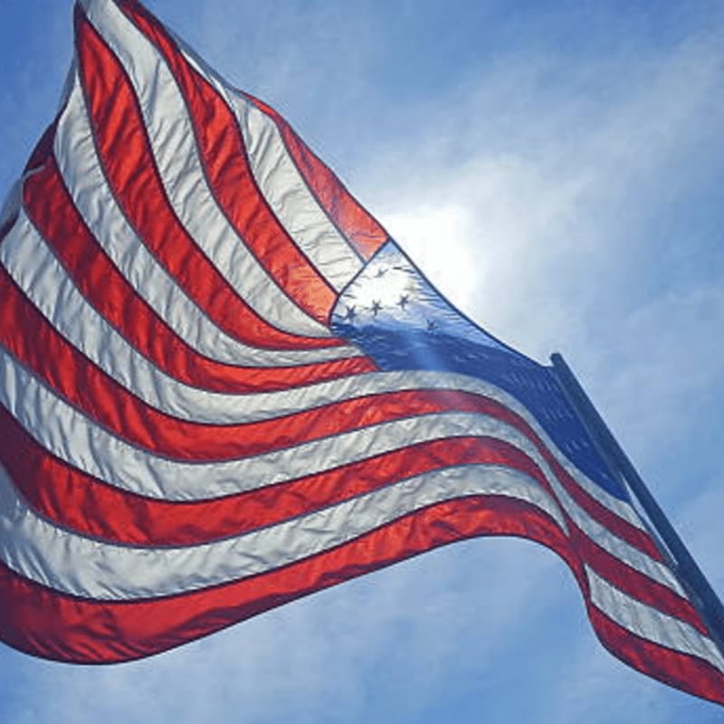 An American flag waves in the foreground against a bright, sunny blue sky.