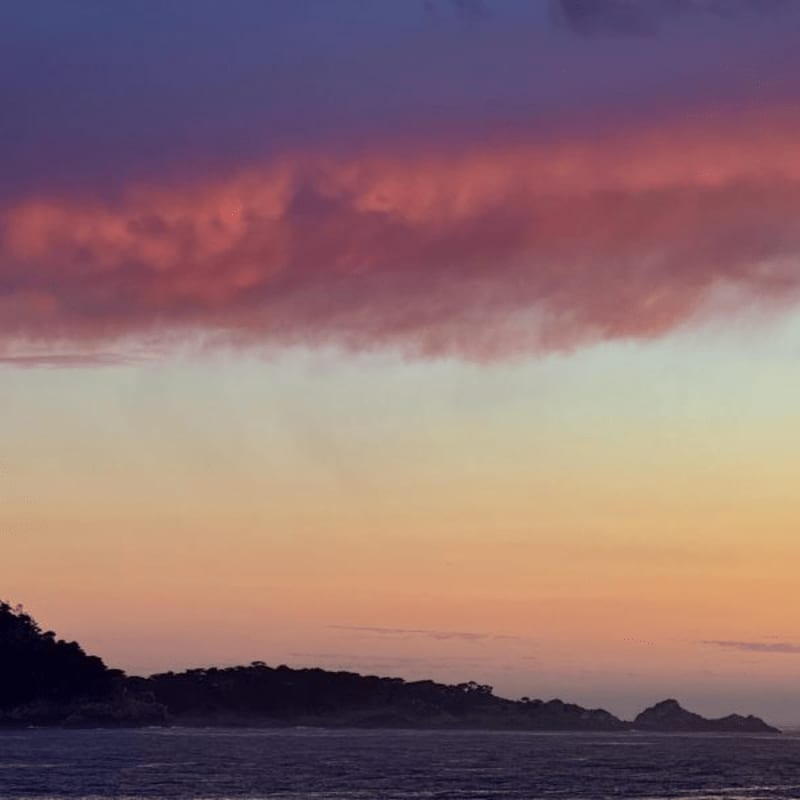 A dark, silhouetted coastline and ocean sit beneath a vibrant sunset sky filled with purple and pink clouds.