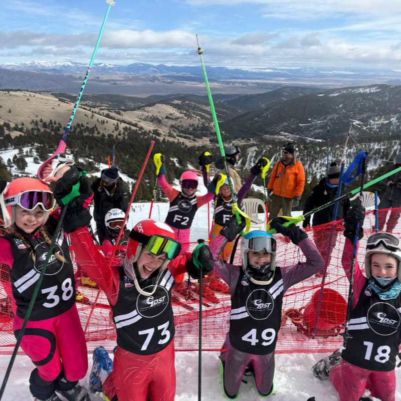 A group of young skiers in racing bibs pose together in the foreground against a backdrop of snowy mountains and a wide valley.