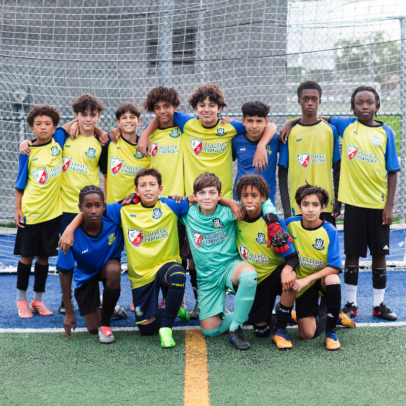 A group of young soccer players in yellow and blue uniforms pose for a photo on a green turf field in front of a goal net.