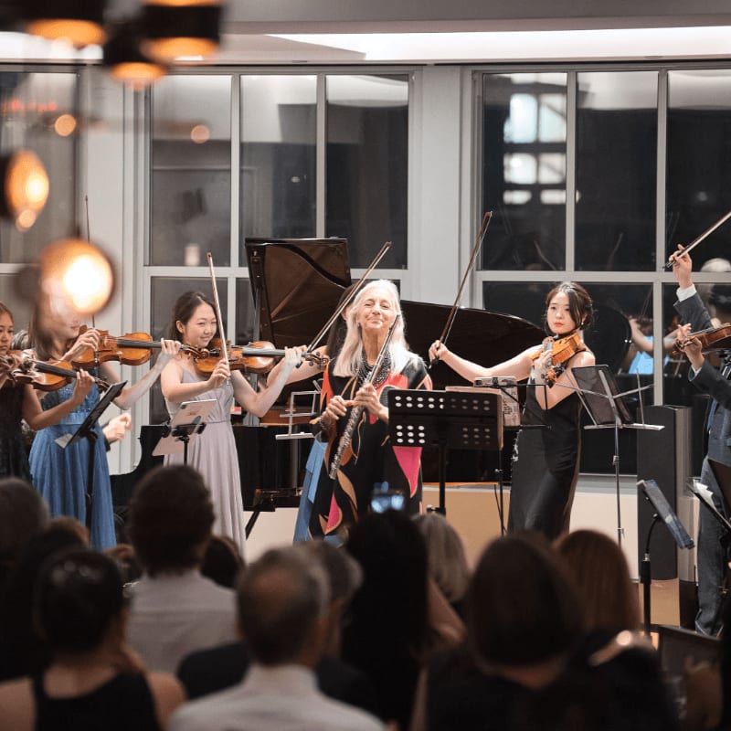 A group of musicians performs on a stage with a grand piano behind them, while an audience watches from the foreground.