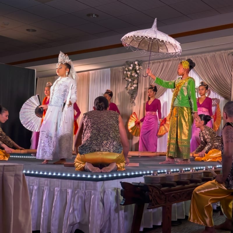 Performers in traditional colorful attire dance on a stage with a white backdrop, while musicians play instruments in the foreground.