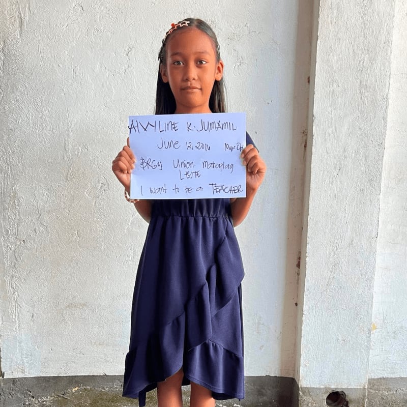 A young girl in a navy blue dress holds a handwritten sign against a plain white wall.