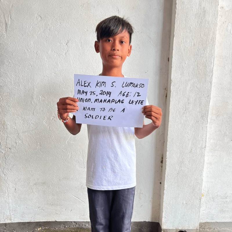 A young boy in a white t-shirt and dark pants holds a handwritten sign against a plain, light-colored wall.