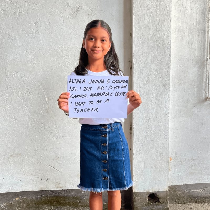 A young girl wearing a white shirt and denim skirt holds a handwritten sign in front of a plain white wall.
