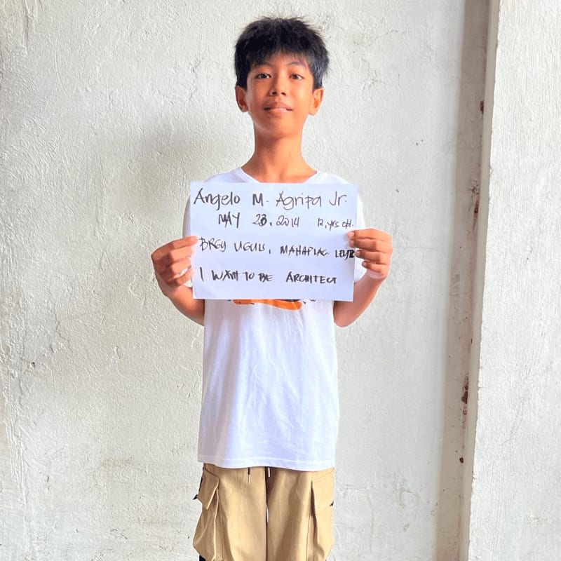 A young boy in a white t-shirt and khaki shorts holds a handwritten sign in front of a plain white wall.