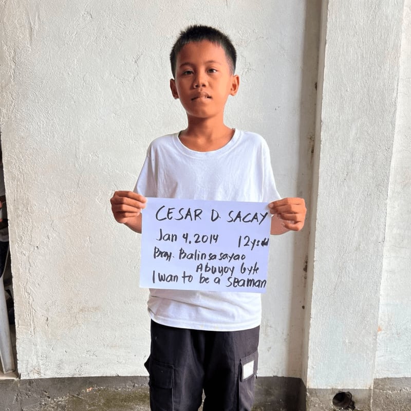A young boy in a white t-shirt and black shorts holds a handwritten sign while standing against a plain white wall.