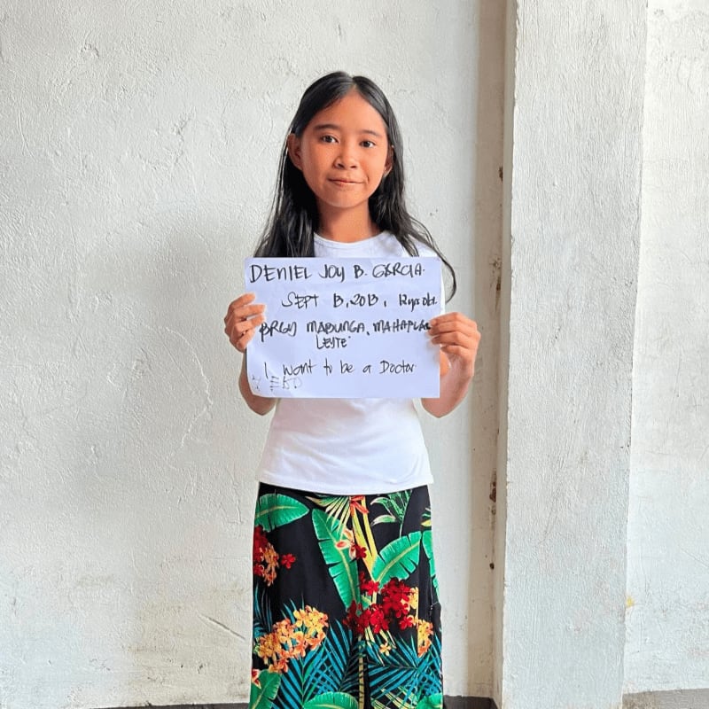 A young girl holding a handwritten sign stands against a plain, light-colored wall.