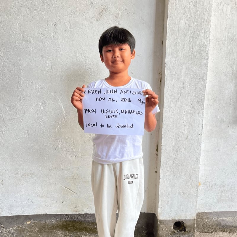 A young boy in a white t-shirt and sweatpants holds a handwritten sign against a plain white wall.