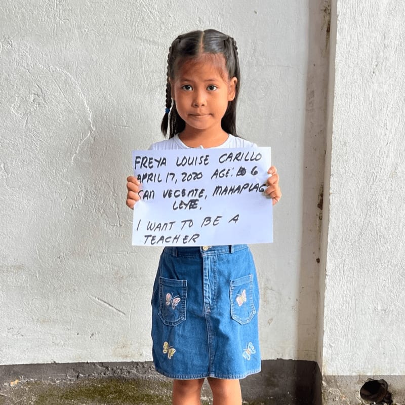 A young girl in a denim skirt holds a sign stating her dream to be a teacher while standing against a plain white wall.