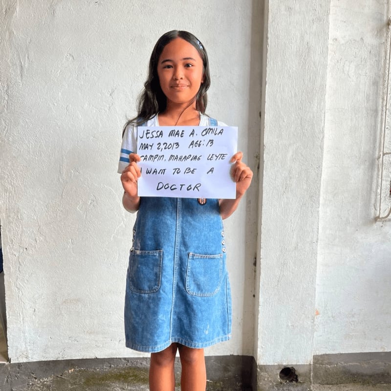 A young girl in a denim dress holds a handwritten sign against a plain white wall.