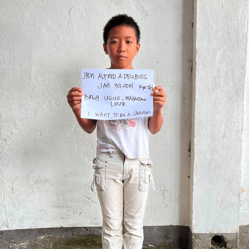 A young boy holds a handwritten sign in front of a plain, textured white wall.