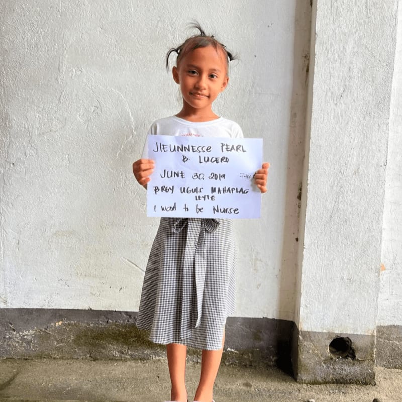 A young girl holds a handwritten sign in front of a plain, textured white wall.