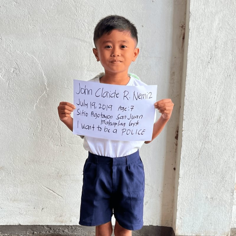 A young boy in a white shirt and blue shorts holds a handwritten sign in front of a plain white wall.